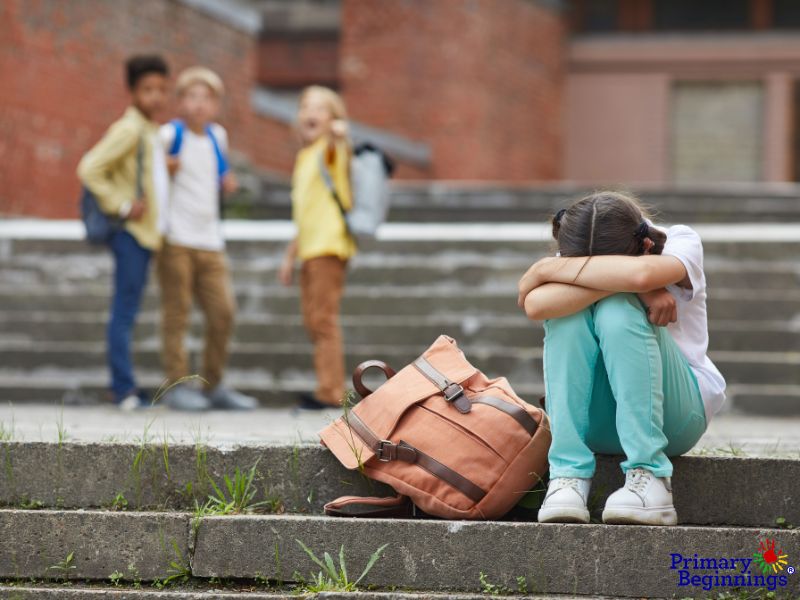 A young girl is sitting on the steps outside of a school while 3 other children are pointing at her. She has experienced bullying in preschool. 